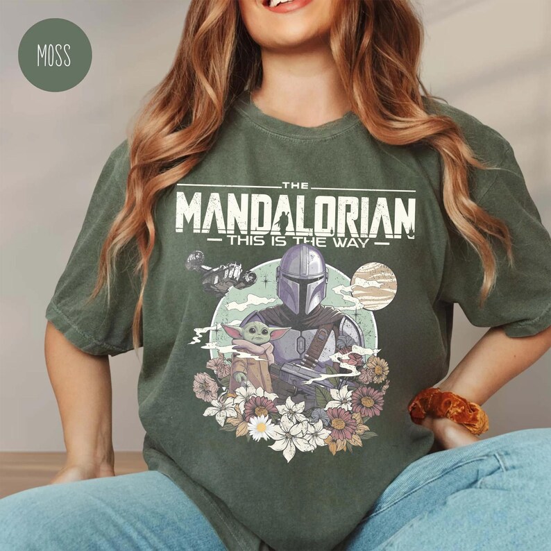 May include: A moss green short-sleeve t-shirt featuring a graphic print of The Mandalorian and Grogu. The design includes the text 'THE MANDALORIAN' and 'THIS IS THE WAY' above the characters, surrounded by a circular background and floral accents.