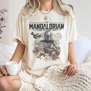 May include: A cream-colored t-shirt featuring a graphic print of The Mandalorian and Grogu. The design includes the text "THE MANDALORIAN THIS IS THE WAY," a spaceship, a planet, and a cluster of flowers in muted tones of brown, pink, and white.