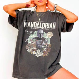 May include: A charcoal gray short-sleeve t-shirt featuring a graphic print of The Mandalorian and Grogu. The text reads THE MANDALORIAN THIS IS THE WAY. The design includes a spaceship, a planet, and a floral arrangement at the bottom of the print.