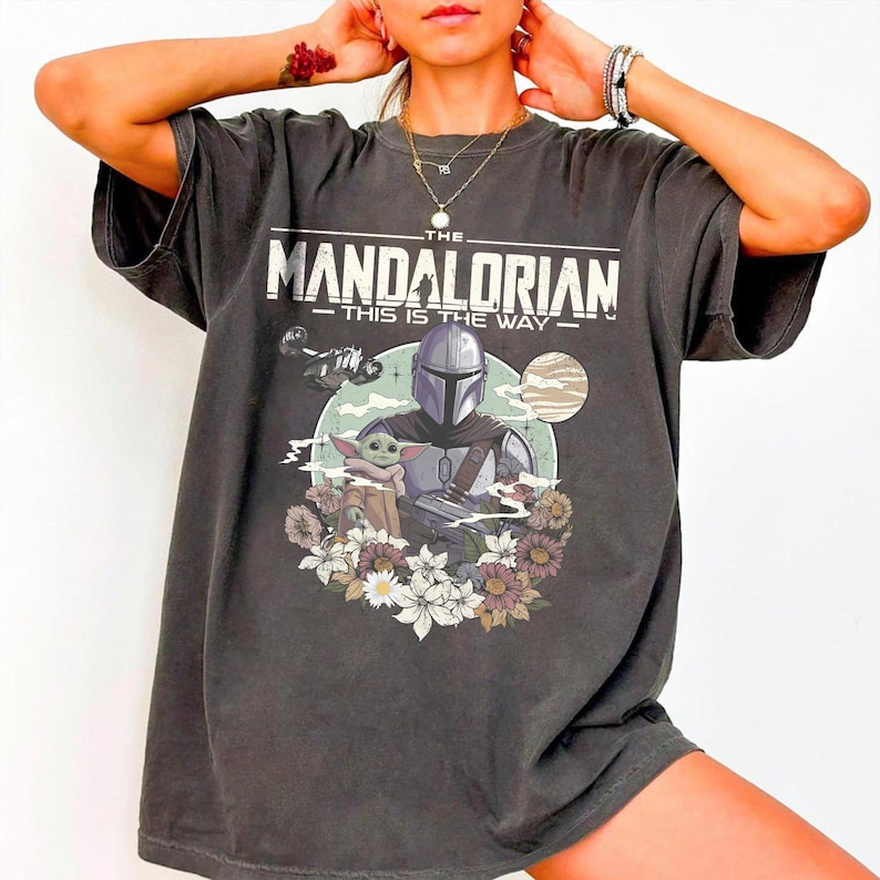 May include: A charcoal gray short-sleeve t-shirt featuring a graphic print of The Mandalorian and Grogu. The text reads THE MANDALORIAN THIS IS THE WAY. The design includes a spaceship, a planet, and a floral arrangement at the bottom of the print.