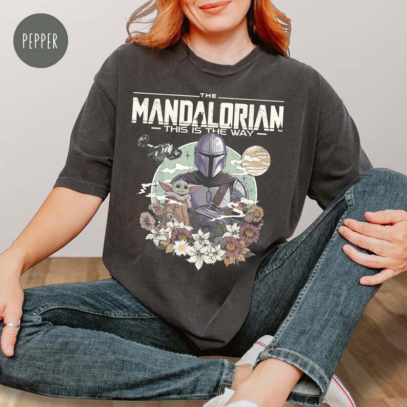 May include: A charcoal gray t-shirt featuring a graphic print of The Mandalorian and Grogu surrounded by flowers. The text reads THE MANDALORIAN THIS IS THE WAY. The design includes a planet and a small spacecraft against a circular background.