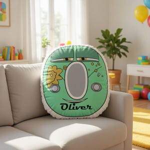 May include: A round, light green decorative pillow featuring a cartoon-style face with sleepy eyes, a large gray oval center, and a yellow flower graphic. The name Oliver is printed in black script at the bottom of the pillow, which has white piping.