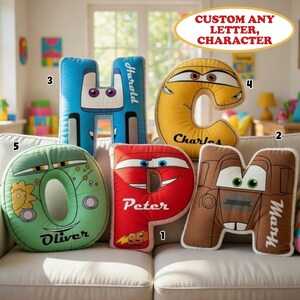 May include: Five plush letter-shaped pillows featuring cartoon car designs with eyes and names printed on them. The pillows are shaped like the letters H, C, O, P, and M, colored blue, yellow, green, red, and brown, with the names Harold, Charles, Oliver, Peter, and Mark.