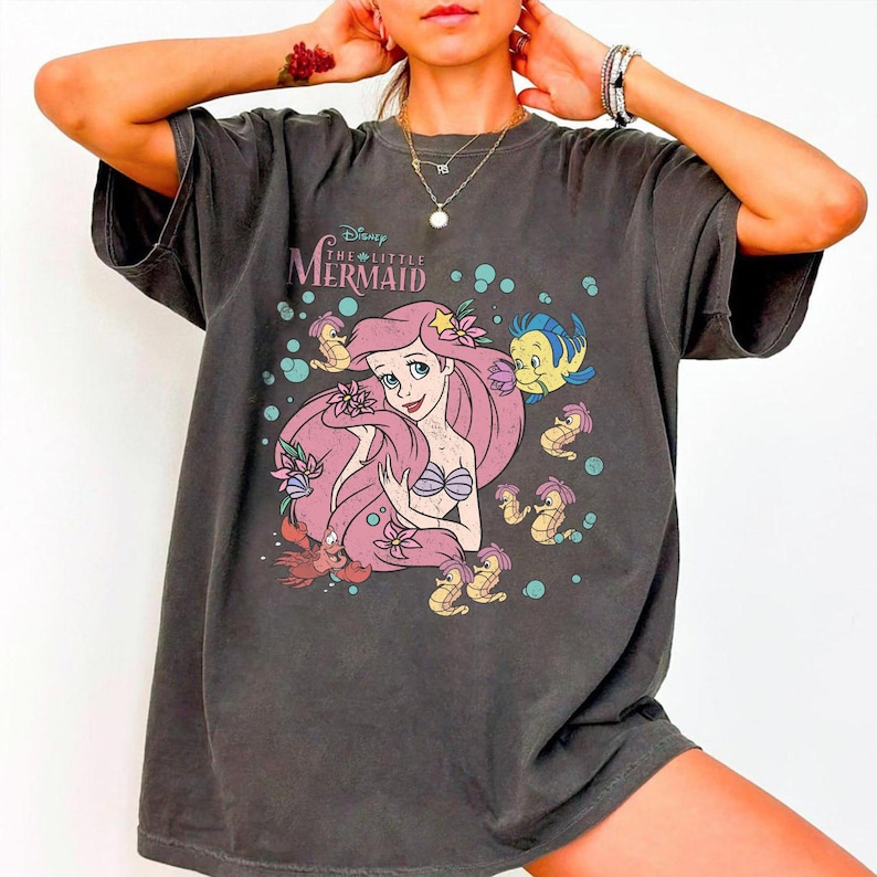 Puede incluir: Una camiseta de manga corta gris carb&oacute;n con un estampado gr&aacute;fico de Ariel de La Sirenita de Disney, rodeada por Flounder, Sebasti&aacute;n y varios caballitos de mar. El dise&ntilde;o incluye burbujas y flores en una paleta de colores apagados.