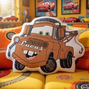 May include: A die-cut throw pillow shaped like the brown tow truck character Mater from the movie Cars. The character features green eyes, a wide smile with buck teeth, and the name Lucas printed in white script on the front hood.