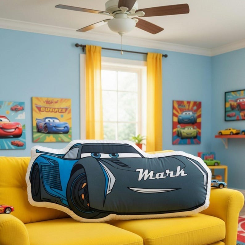 May include: A plush pillow shaped like a blue race car with black wheels and a white spoiler. The car features blue eyes on the windshield and the name Mark printed in white script on the side. The pillow is resting on a yellow sofa.