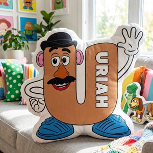 Puede incluir: Un gran coj&iacute;n blanco en forma de U con un dise&ntilde;o de personaje de dibujos animados. El personaje tiene una cara marr&oacute;n, un bigote negro y un sombrero negro. El nombre "URIAH" est&aacute; impreso verticalmente. Zapatos azules y una mano que saluda completan el dise&ntilde;o.