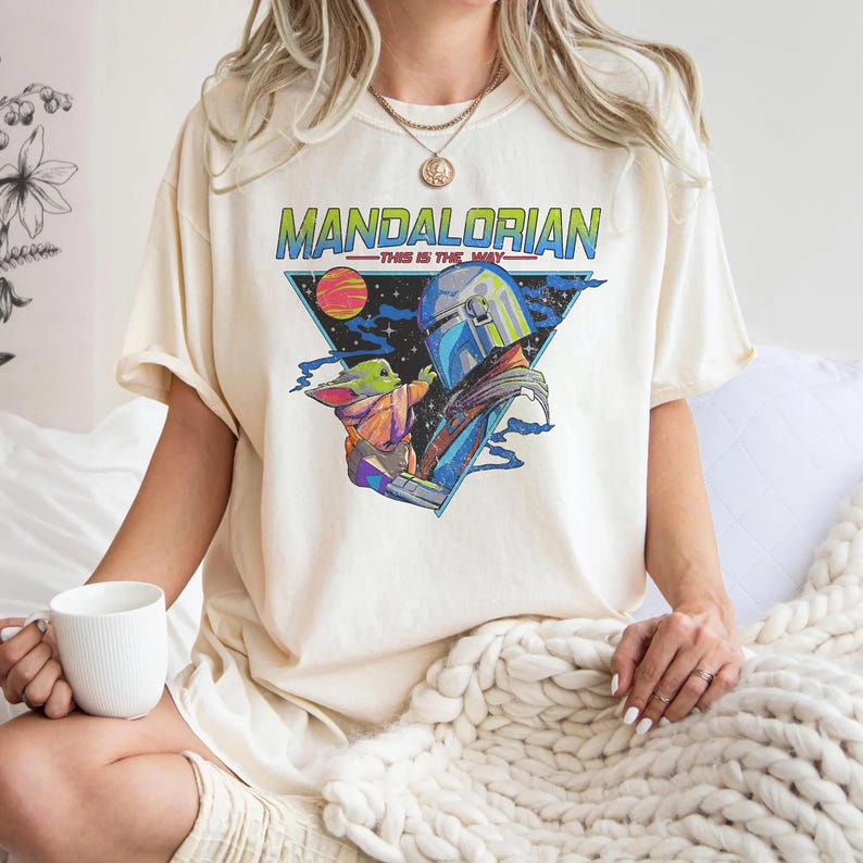 May include: A cream-colored short-sleeve t-shirt featuring a graphic print of The Mandalorian and Grogu. The design includes the text "MANDALORIAN" and "THIS IS THE WAY" in a colorful, retro-inspired style set against a dark triangular background.