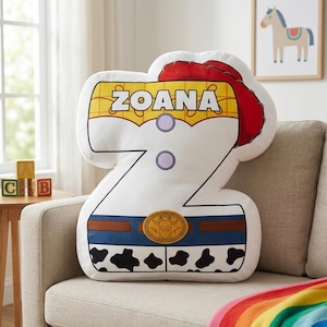 Puede incluir: Un coj&iacute;n decorativo con forma de la letra "Z" con un dise&ntilde;o que se asemeja a un atuendo de vaquera. El coj&iacute;n presenta una parte superior amarilla y blanca con el nombre "ZOANA", un sombrero rojo y un cintur&oacute;n azul con una hebilla dorada. La parte inferior tiene un patr&oacute;n de piel de vaca en blanco y negro.