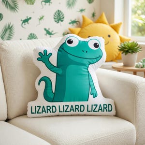May include: A white, lizard-shaped pillow with a teal green lizard illustration waving its hand. The pillow has the word "LIZARD" printed three times in green. A yellow sun-shaped pillow is in the background.
