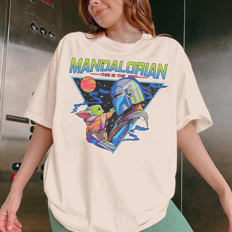 May include: A cream-colored short-sleeve t-shirt featuring a graphic print of The Mandalorian and Grogu. The design includes a blue triangle, a planet, and the text "MANDALORIAN" in green and "THIS IS THE WAY" in red against a starry background.