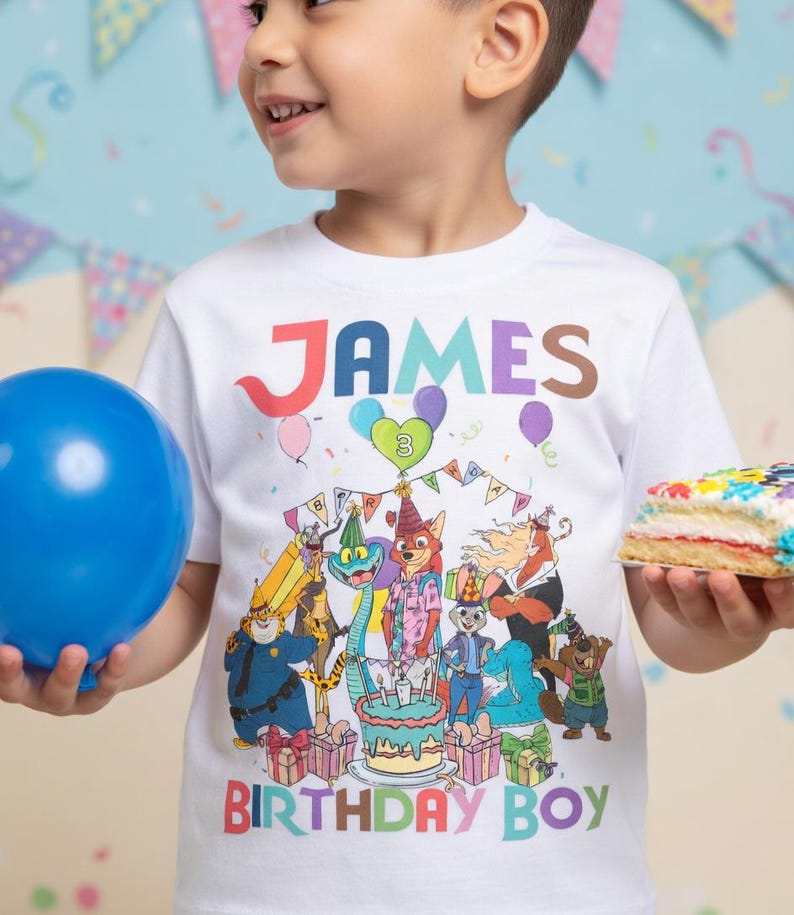 Pode incluir: Camiseta branca com o nome "JAMES" em letras coloridas e a mensagem "BIRTHDAY BOY". A camiseta apresenta uma ilustra&ccedil;&atilde;o de desenho animado de animais em volta de um bolo de anivers&aacute;rio. Um bal&atilde;o azul e um peda&ccedil;o de bolo tamb&eacute;m s&atilde;o vis&iacute;veis.