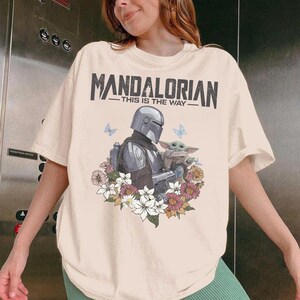 May include: Cream t-shirt with a graphic of The Mandalorian and Grogu surrounded by flowers and butterflies. The text "MANDALORIAN - THIS IS THE WAY" is printed across the top in black.