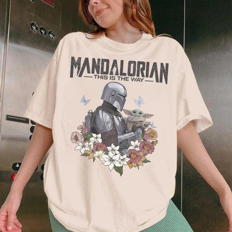 May include: Cream t-shirt with a graphic of The Mandalorian and Grogu surrounded by flowers and butterflies. The text "MANDALORIAN - THIS IS THE WAY" is printed across the top in black.