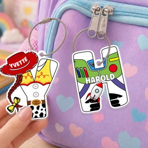 May include: Two custom acrylic letter keychains attached to a purple heart-patterned backpack. One is a letter Y shaped like a cowgirl outfit with a red hat labeled YVETTE. The other is a letter H shaped like a space ranger suit labeled HAROLD.
