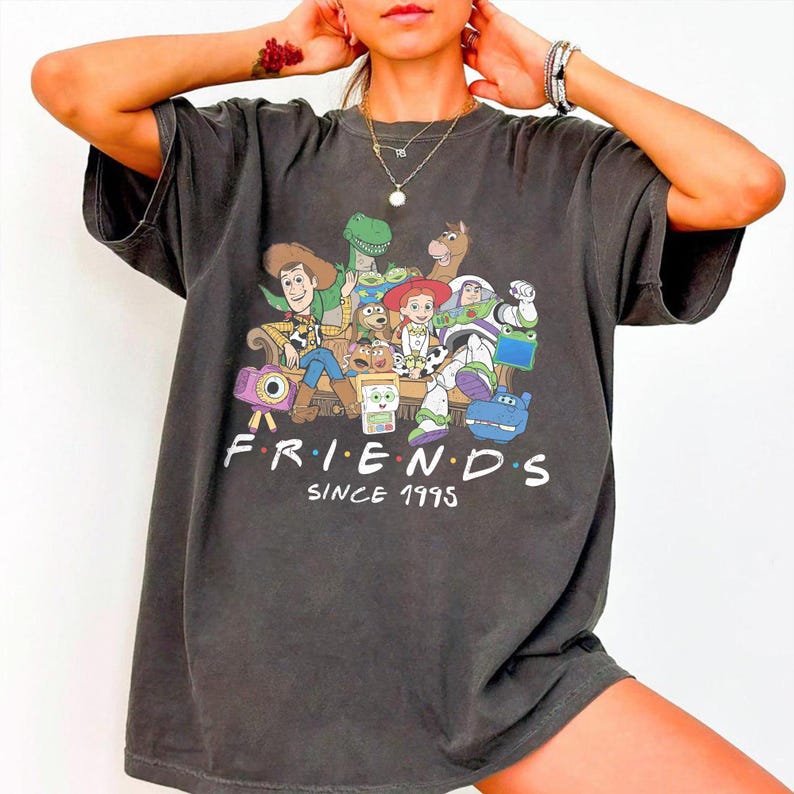 Puede incluir: Una camiseta de manga corta gris carb&oacute;n con un estampado gr&aacute;fico de varios personajes animados sentados juntos. Debajo de los personajes, el texto dice "FRIENDS" en fuente blanca con puntos de colores, seguido de "SINCE 1995".