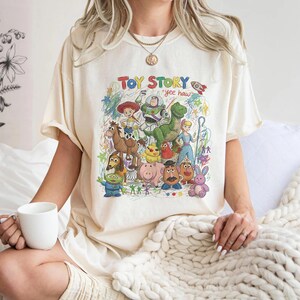 K&ouml;nnte beinhalten: Ein cremefarbenes T-Shirt mit einem bunten Grafikdruck von Toy Story-Figuren wie Woody, Buzz Lightyear, Rex und anderen. Das Design enth&auml;lt den Text "TOY STORY" und "yee haw" in einem handgezeichneten, buntstiftartigen k&uuml;nstlerischen Stil.