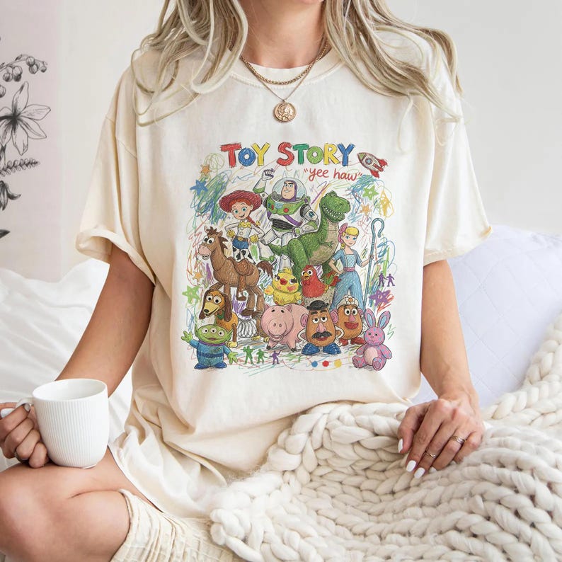 K&ouml;nnte beinhalten: Ein cremefarbenes T-Shirt mit einem bunten Grafikdruck von Toy Story-Figuren wie Woody, Buzz Lightyear, Rex und anderen. Das Design enth&auml;lt den Text "TOY STORY" und "yee haw" in einem handgezeichneten, buntstiftartigen k&uuml;nstlerischen Stil.