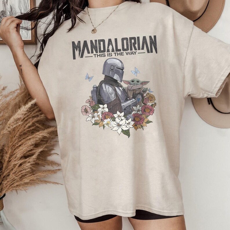 May include: Beige t-shirt with a graphic of The Mandalorian and Grogu, surrounded by flowers and butterflies. The text "MANDALORIAN - THIS IS THE WAY" is printed across the top in black.