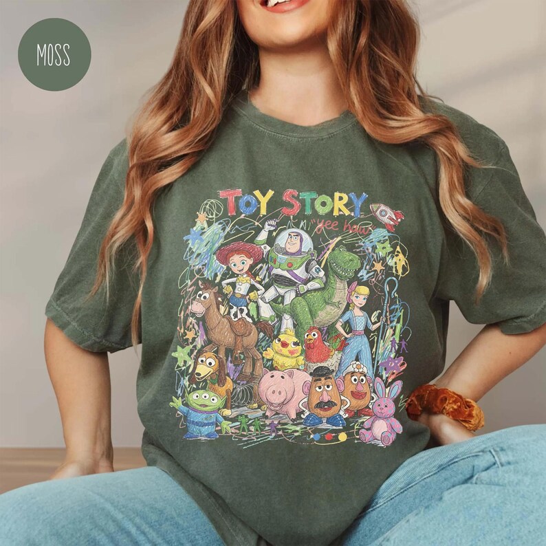 K&ouml;nnte beinhalten: Ein moosgr&uuml;nes Kurzarm-T-Shirt mit einem farbenfrohen Grafikdruck der Toy Story-Figuren wie Woody, Buzz Lightyear, Jessie, Rex und anderen. Das Design enth&auml;lt den Text "TOY STORY" und "yee haw" mit krakeligen Akzenten.