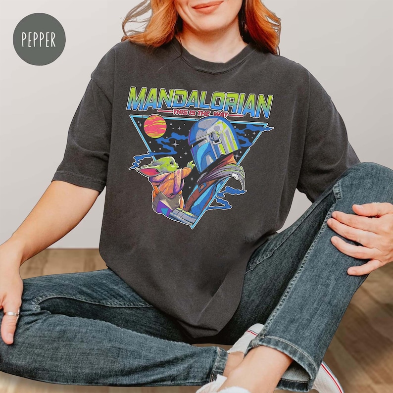 May include: A charcoal gray short-sleeve t-shirt featuring a graphic print of The Mandalorian and Grogu. The design includes the text "MANDALORIAN" and "THIS IS THE WAY" in blue and green, set against a triangular background with a planet.