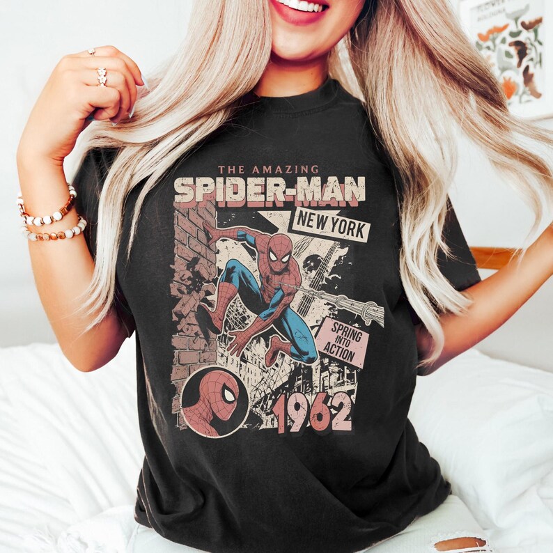 Op de afbeelding: Zwart T-shirt met een afbeelding van Spider-Man in actie, met de tekst "THE AMAZING SPIDER-MAN NEW YORK" en "SPRING INTO ACTION 1962". Het ontwerp bevat een bakstenen muur en een stadsgezicht.