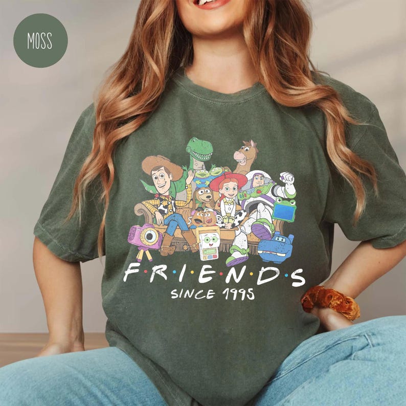 Puede incluir: Una camiseta de manga corta color verde musgo con un estampado gr&aacute;fico de varios personajes animados sentados en un sof&aacute;. Debajo de los personajes, el texto "FRIENDS SINCE 1995" aparece en color blanco. La camiseta tiene cuello redondo y corte holgado.