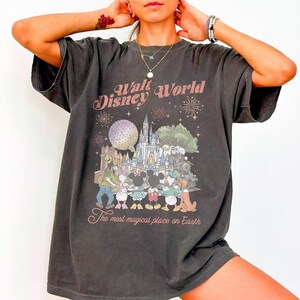 May include: Dark gray t-shirt featuring a vintage-style graphic of Walt Disney World. The design includes the park's castle, characters, and the text "The most magical place on Earth."
