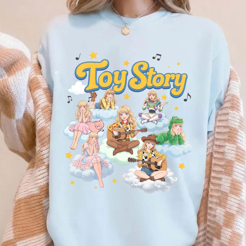 May include: A light blue sweatshirt featuring a graphic print with the text Toy Story in yellow. The design includes multiple blonde characters in various outfits sitting on white clouds, surrounded by yellow stars and black musical notes.