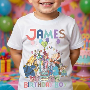 Pode incluir: Camiseta branca com o nome "JAMES" em letras coloridas. A camiseta apresenta uma ilustra&ccedil;&atilde;o de desenho animado de animais celebrando um anivers&aacute;rio, com o texto "BIRTHAY BOY". Bal&otilde;es e um bolo tamb&eacute;m s&atilde;o vis&iacute;veis.