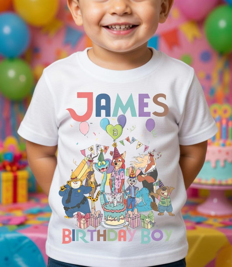 Pode incluir: Camiseta branca com o nome "JAMES" em letras coloridas. A camiseta apresenta uma ilustra&ccedil;&atilde;o de desenho animado de animais celebrando um anivers&aacute;rio, com o texto "BIRTHAY BOY". Bal&otilde;es e um bolo tamb&eacute;m s&atilde;o vis&iacute;veis.