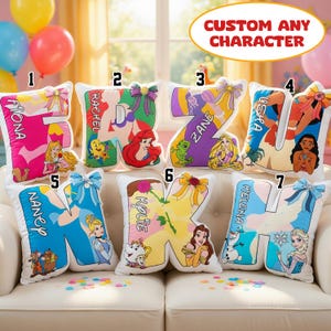 May include: Seven letter-shaped scatter cushions featuring various cartoon characters and names. Each cushion is decorated with a colourful bow and displays a unique character theme, including princesses and sidekicks, with custom names printed on each.