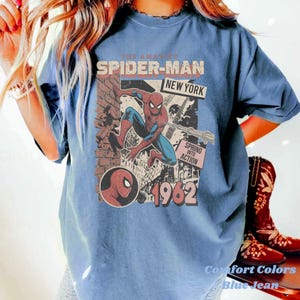 Op de afbeelding: Blauw Comfort Colors T-shirt met een vintage Spider-Man graphic. Het ontwerp bevat Spider-Man in actie, de tekst "The Amazing Spider-Man New York" en het jaar "1962". Het shirt heeft de kleur Blue Jean.