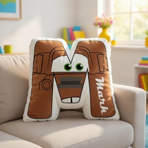 May include: A letter M shaped throw pillow featuring a brown and white design inspired by a tow truck character with green eyes and a front grille. The name Mark is printed in white vertical text on the right side of the letter shaped cushion.
