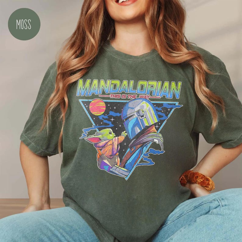 May include: A moss green short-sleeve t-shirt featuring a graphic print of The Mandalorian and Grogu. The design includes the text "MANDALORIAN" and "THIS IS THE WAY" in a retro font, set against a triangular space background with a planet.