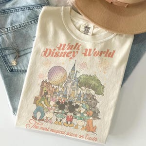 May include: Cream-colored t-shirt with a graphic of Disney World characters and the text "Walt Disney World" and "The most magical place on Earth." The shirt is folded on top of blue jeans, with a necklace and a tan hat.