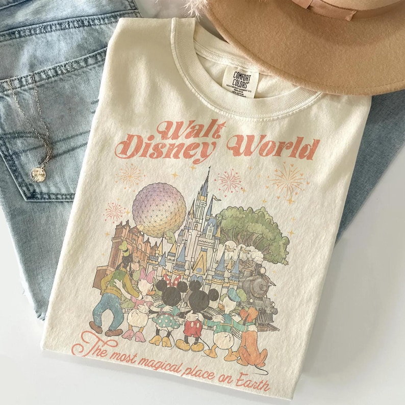 May include: Cream-colored t-shirt with a graphic of Disney World characters and the text "Walt Disney World" and "The most magical place on Earth." The shirt is folded on top of blue jeans, with a necklace and a tan hat.