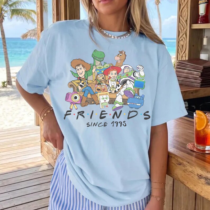 Puede incluir: Una camiseta azul claro con un estampado gr&aacute;fico de varios personajes de juguetes animados sentados juntos. Debajo de los personajes, el texto "FRIENDS SINCE 1995" aparece en letras negras. La camiseta tiene cuello redondo y mangas cortas.