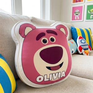 Puede incluir: Un coj&iacute;n rosa con forma de oso con un borde blanco y el nombre "OLIVIA" en texto blanco. El coj&iacute;n tiene una cara sonriente con ojos marrones oscuros y una nariz y boca de color rojo oscuro. Otros cojines decorativos son visibles en el fondo.