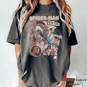 Op de afbeelding: Donkergrijze t-shirt met een vintage Spider-Man graphic. Het ontwerp bevat de tekst "THE AMAZING SPIDER-MAN NEW YORK" en "1962". De graphic toont Spider-Man in actie, met een bakstenen muur achtergrond. Het shirt is een Comfort Colors Pepper.