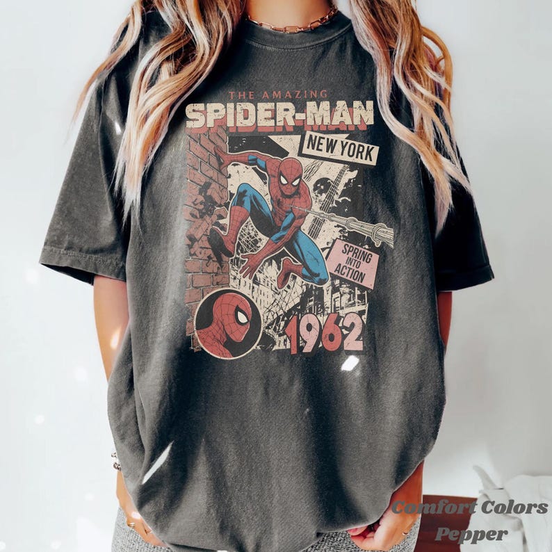 Op de afbeelding: Donkergrijze t-shirt met een vintage Spider-Man graphic. Het ontwerp bevat de tekst "THE AMAZING SPIDER-MAN NEW YORK" en "1962". De graphic toont Spider-Man in actie, met een bakstenen muur achtergrond. Het shirt is een Comfort Colors Pepper.