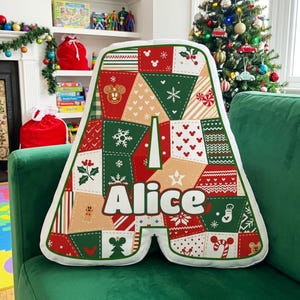 May include: A letter-shaped throw pillow in the form of the letter A, featuring a patchwork design of red, green, and beige holiday patterns. The fabric displays Mickey Mouse icons, snowflakes, holly, and the name Alice printed in white letters.