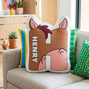 Puede incluir: Un coj&iacute;n decorativo con forma de la letra "H" con un dise&ntilde;o de cara de caballo. El coj&iacute;n es marr&oacute;n, rosa y blanco, con el nombre "HENRY" impreso verticalmente en blanco. Se ven otros cojines amarillos, verdes y blancos.