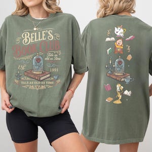 May include: Olive green t-shirt with "Belle's Book Club" graphic. The front design features a rose in a glass dome, books, and text. The back has a similar design with characters and books. "Tale as old as time" is also printed on the shirt.