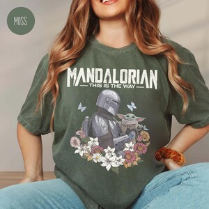 May include: Moss green t-shirt with the text "MANDALORIAN - THIS IS THE WAY" above an illustration of the Mandalorian and Grogu surrounded by flowers and butterflies. The shirt has a relaxed fit.