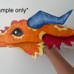 Puede incluir: Una colorida máscara de cabeza de dragón con detalles en naranja, amarillo, rojo y azul. La cabeza del dragón está pintada con detalles intrincados, incluyendo un ojo grande y patrones decorativos. La máscara es sostenida por el brazo de una persona.