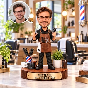 Op de afbeelding: Een gepersonaliseerde bobblehead-figuur van een kapper met bruin haar en een bril, gekleed in een zwart overhemd en bruin leren schort. De figuur houdt een schaar en tondeuse vast op een houten voet met het plaatje MICHAEL BEST BARBER EVER.