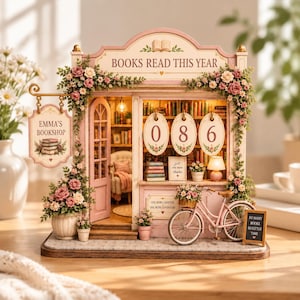 May include: A miniature bookshop model titled Emma's Bookshop with a Books Read This Year sign, featuring a pink bicycle, floral decorations, and three oval plaques displaying the number 086. The interior shows shelves of books and a chair.