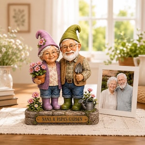 May include: A figurine of two elderly gnomes standing on a stone base labeled NANA & PAPA'S GARDEN. The female gnome wears a purple hat and holds a potted flower, while the male gnome wears a green hat and holds a trowel. A photo frame sits nearby.