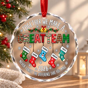 Peut inclure: Une décoration de Noël ronde en verre transparent à facettes suspendue par un ruban rouge. Elle porte l'inscription TOGETHER WE MAKE A GREAT TEAM et NEVER FORGET THE DIFFERENCE THAT YOU MAKE, avec cinq chaussettes nommées Isabella, Sophie, Thomas, Martin et Daisy, ainsi que l'année 2026.